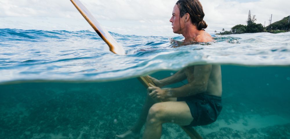 A person with long hair and wearing dark shorts floats on a surfboard in clear blue ocean water. The horizon shows a partly cloudy sky and distant trees, conveying a serene and adventurous mood.