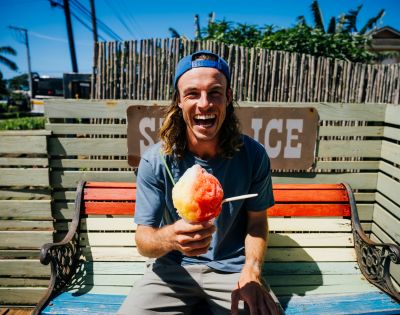 A cheerful person with long hair and a backward hat sits on a bench, joyfully holding a colorful shaved ice under a bright blue sky.