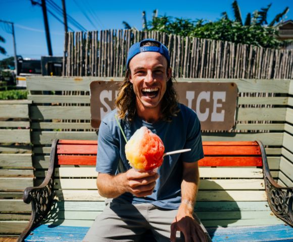 A cheerful person with long hair and a backward hat sits on a bench, joyfully holding a colorful shaved ice under a bright blue sky.