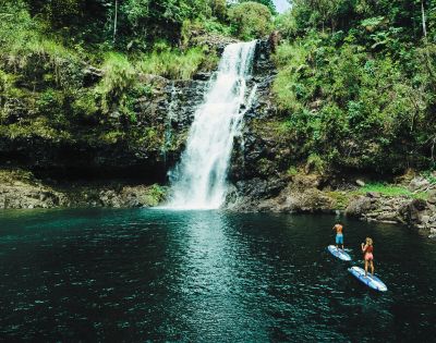 Two people paddleboard on a serene, emerald pool in front of a cascading waterfall surrounded by lush green foliage and rocky cliffs, conveying tranquility.