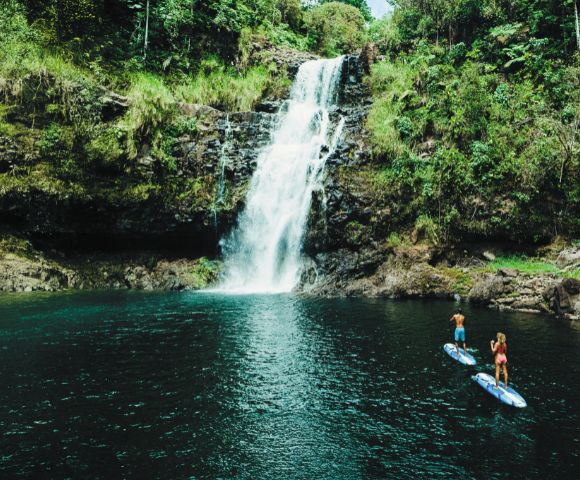 Two people paddleboard on a serene, emerald pool in front of a cascading waterfall surrounded by lush green foliage and rocky cliffs, conveying tranquility.