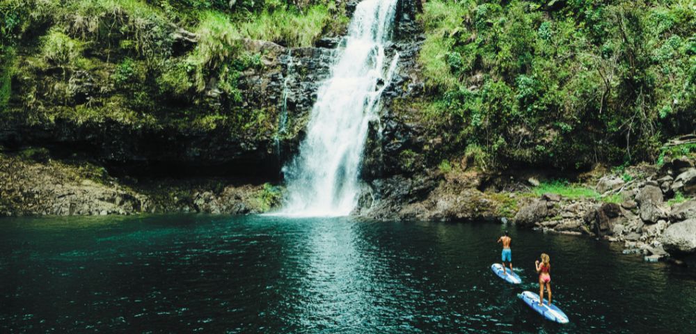 Two people paddleboarding on serene blue water in front of a lush, green forest with a cascading waterfall. The scene exudes tranquility and adventure.