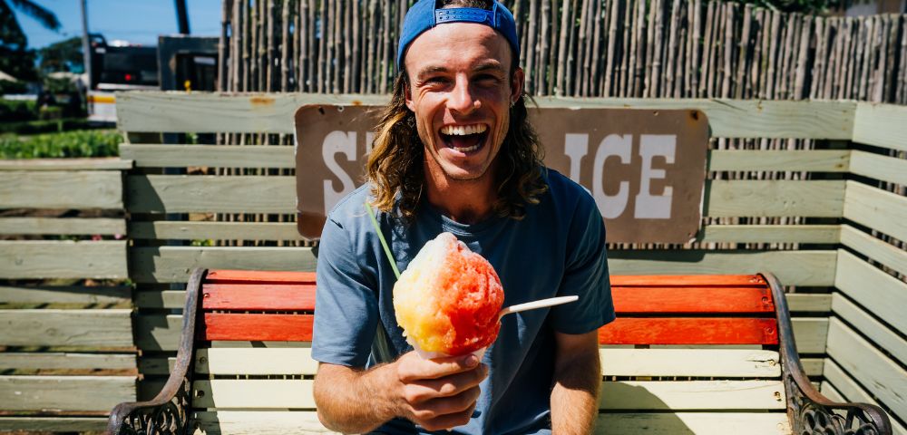 A man with long hair and a backward cap sits on a bench, laughing and holding a colorful shaved ice. The scene is bright and cheerful, suggesting a sunny day.