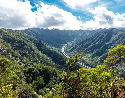 Scenic mountain landscape with a winding road cutting through lush green valleys, under a partly cloudy sky. The view conveys tranquility and natural beauty.