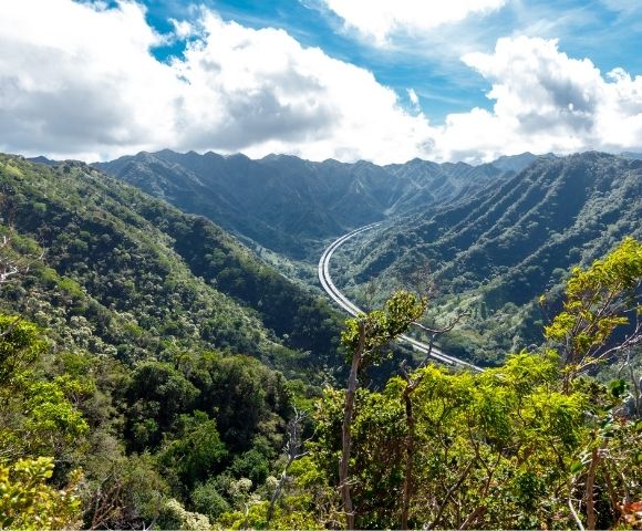 Scenic mountain landscape with a winding road cutting through lush green valleys, under a partly cloudy sky. The view conveys tranquility and natural beauty.