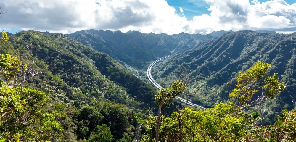 Scenic mountain landscape with a winding road cutting through lush green valleys, under a partly cloudy sky. The view conveys tranquility and natural beauty.