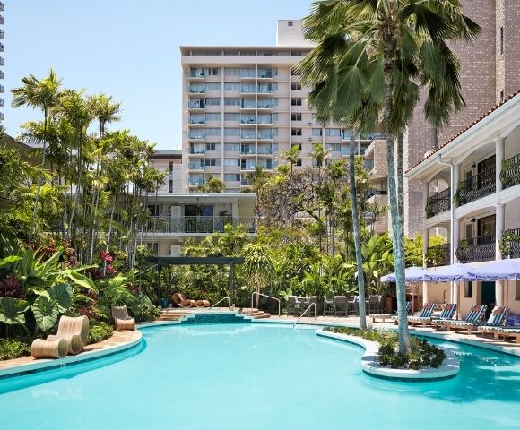 Luxurious pool area surrounded by tropical plants and palm trees, with lounge chairs and umbrellas. A modern hotel building is visible in the background.
