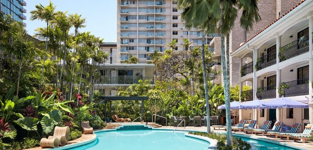 Luxurious pool area surrounded by tropical plants and palm trees, with lounge chairs and umbrellas. A modern hotel building is visible in the background.