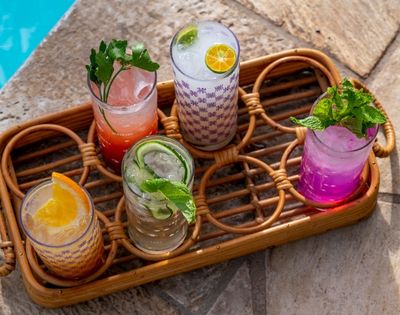 Colorful cocktails in decorated glasses sit on a wicker tray by a pool. Garnished with fresh mint, citrus, and cucumber, evoking a refreshing summer vibe.