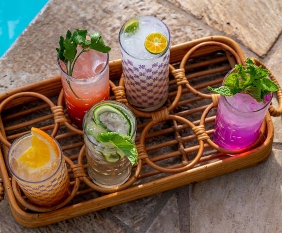 Colorful cocktails in decorated glasses sit on a wicker tray by a pool. Garnished with fresh mint, citrus, and cucumber, evoking a refreshing summer vibe.