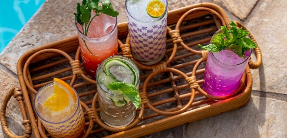 Colorful cocktails in decorated glasses sit on a wicker tray by a pool. Garnished with fresh mint, citrus, and cucumber, evoking a refreshing summer vibe.