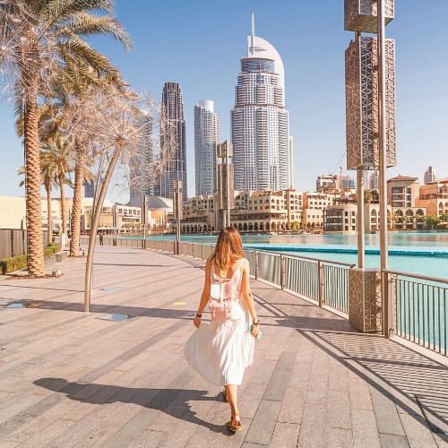 A woman in a white dress walks along a sunny waterfront promenade in a cityscape with modern skyscrapers, palm trees, and a clear blue sky.