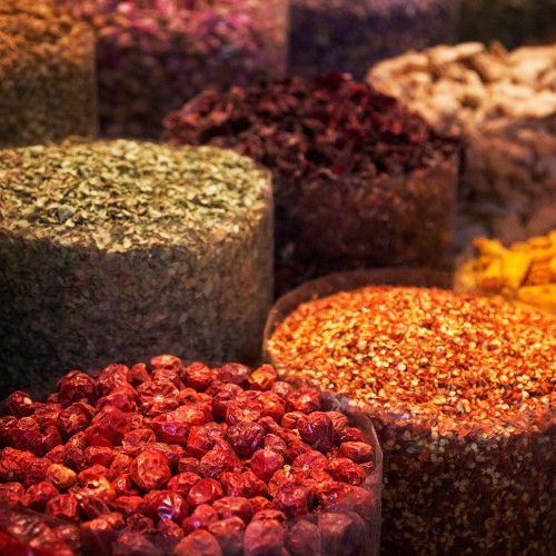 Assorted spices in large, colorful baskets at a market. Red chilies, green herbs, and orange flakes create a vibrant, aromatic display.