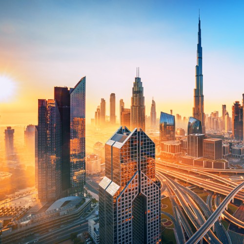 A cityscape of Dubai at sunset, featuring tall skyscrapers, including the Burj Khalifa, with glowing sunlight reflecting off the buildings and roads.