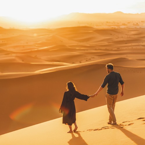 A couple holding hands walks on sunlit desert dunes at sunset. The warm glow casts long shadows, evoking romance and tranquility in a vast landscape.