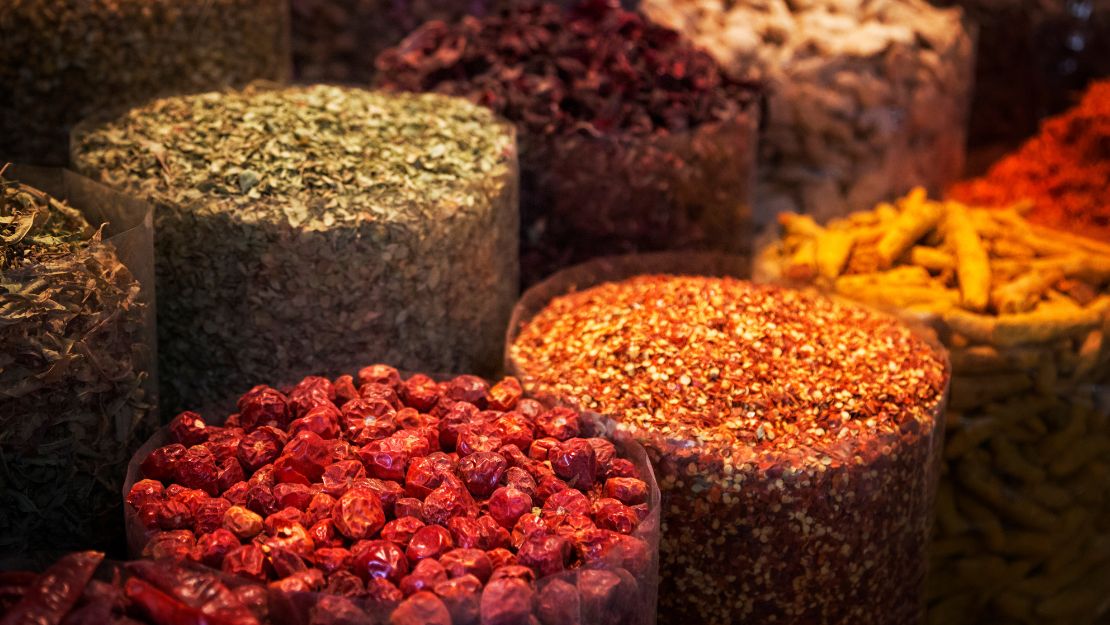 Assorted spices in large, colorful baskets at a market. Red chilies, green herbs, and orange flakes create a vibrant, aromatic display.