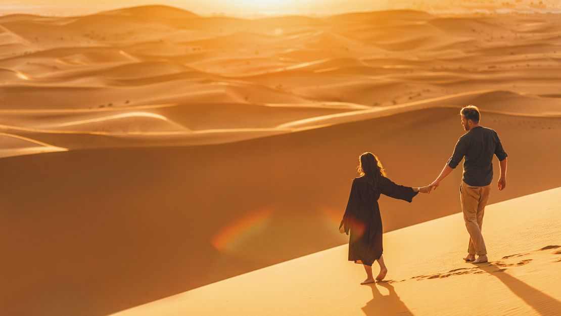 A couple holding hands walks on sunlit desert dunes at sunset. The warm glow casts long shadows, evoking romance and tranquility in a vast landscape.