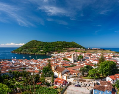 Coastal town with red-roofed buildings nestled by a blue ocean. Lush green island in the background under a clear blue sky.