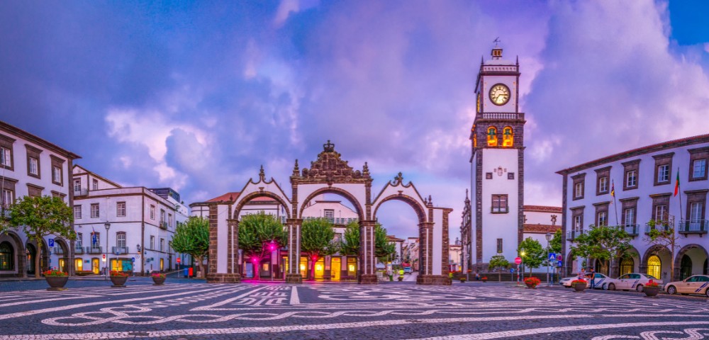 A scenic plaza at sunset with ornate arches and a clock tower. The patterned cobblestones and glow from colorful lights create a calm, picturesque ambiance.