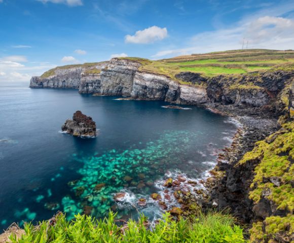Coastal cliff view with clear turquoise waters surrounds rocky outcrops. Green fields top the cliffs, under a blue sky with scattered white clouds. Serene and picturesque.
