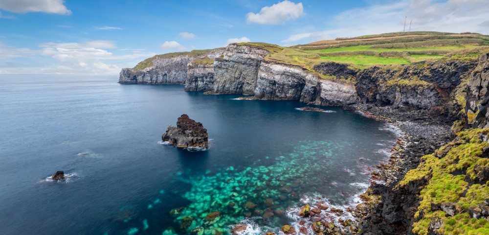 Coastal cliff view with clear turquoise waters surrounds rocky outcrops. Green fields top the cliffs, under a blue sky with scattered white clouds. Serene and picturesque.