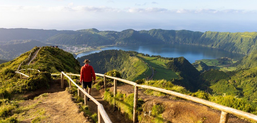 A person in a red jacket walks down a dirt path lined with wooden posts, overlooking a lush green landscape and a serene blue lake under a clear sky.