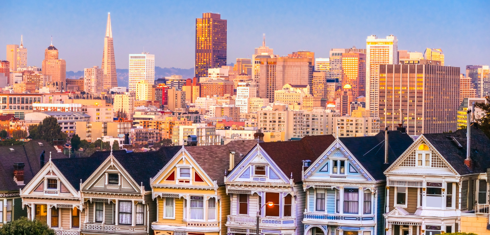 Colorful Victorian houses, known as the Painted Ladies, stand in the foreground with the San Francisco skyline glowing at sunset in the background.
