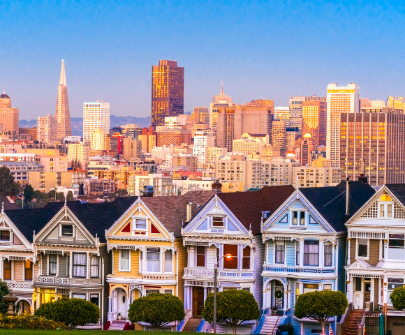 Colorful Victorian houses, known as the Painted Ladies, stand in the foreground with the San Francisco skyline glowing at sunset in the background.