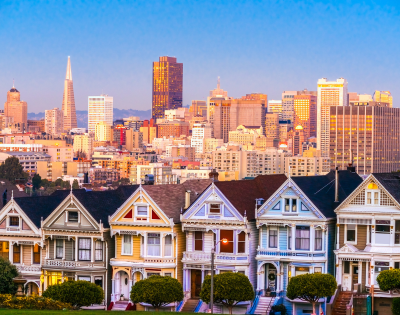 Colorful Victorian houses, known as the Painted Ladies, stand in the foreground with the San Francisco skyline glowing at sunset in the background.