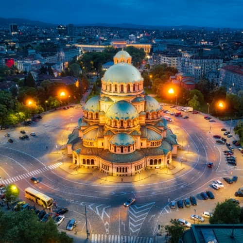 Aerial view of a brightly lit cathedral with green domes in a circular plaza at dusk, surrounded by city buildings and glowing streetlights, conveying tranquility.