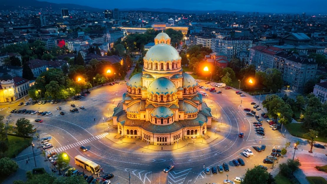 Aerial view of a brightly lit cathedral with green domes in a circular plaza at dusk, surrounded by city buildings and glowing streetlights, conveying tranquility.