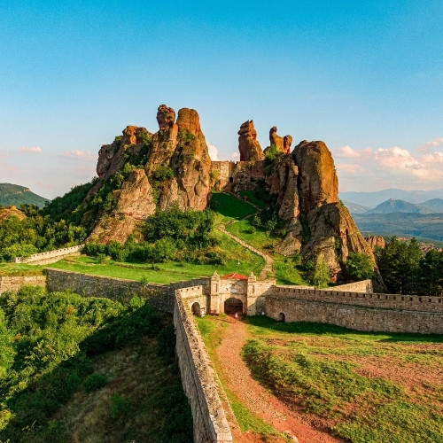 Majestic rock formations rise behind an ancient fortress, surrounded by lush greenery. A long stone wall extends, with mountains under a bright blue sky.