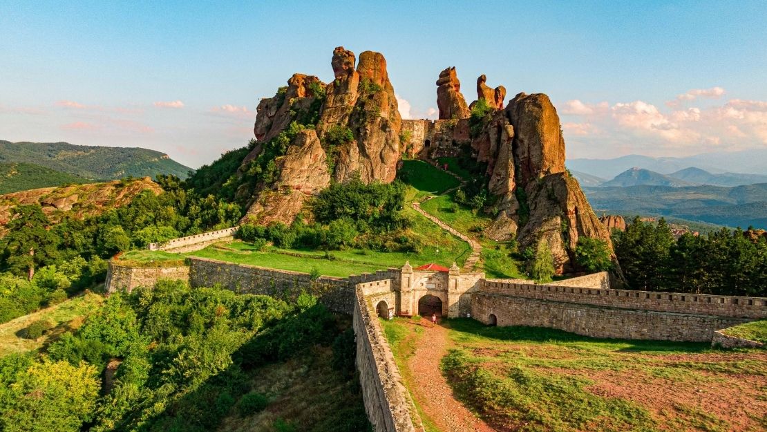 Majestic rock formations rise behind an ancient fortress, surrounded by lush greenery. A long stone wall extends, with mountains under a bright blue sky.