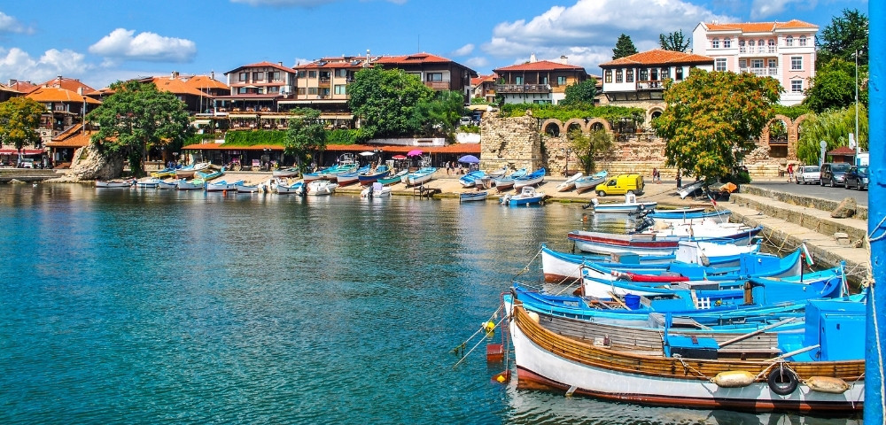Coastal scene with colorful wooden boats docked in a calm blue harbor. Historic buildings and lush greenery line the shore, under a sky with fluffy clouds.
