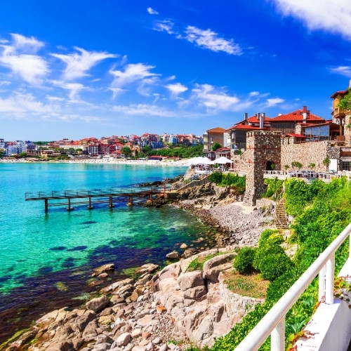 Scenic seaside pathway with stone retaining walls and red-roofed buildings, overlooking turquoise waters and a vibrant town under a clear blue sky.