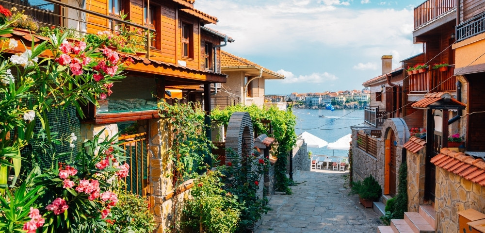 Narrow stone street lined with colorful flowers and wooden houses leads to a view of the sea under a bright sky, creating a peaceful and inviting scene.