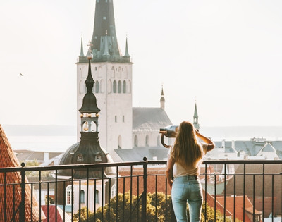 A woman with long hair looks through binoculars from a balcony, facing historic stone towers with tall spires.