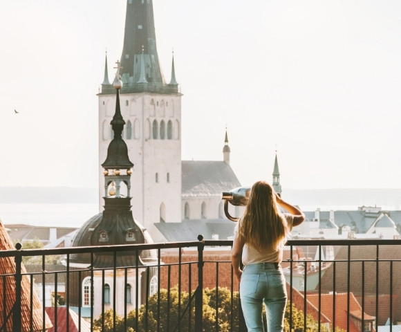 A woman with long hair looks through binoculars from a balcony, facing historic stone towers with tall spires.