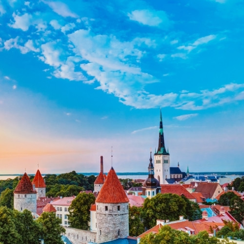 A scenic view of a historic cityscape at sunset, featuring medieval stone towers with red roofs, lush greenery, and a tall church spire under a bright blue sky.