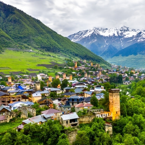Vibrant valley town with medieval towers nestled in lush green hills, framed by majestic snow-capped mountains under a cloudy sky.