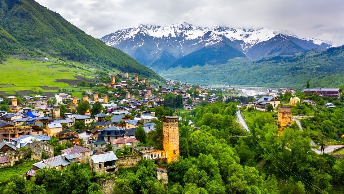 Vibrant valley town with medieval towers nestled in lush green hills, framed by majestic snow-capped mountains under a cloudy sky.