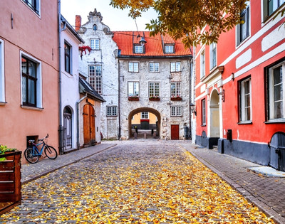 A picturesque cobblestone street lined with colorful buildings in autumn. Leaves scatter the ground, and a bicycle rests on the left. A charming archway leads to more buildings beyond.