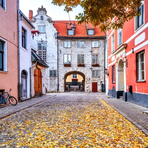 A picturesque cobblestone street lined with colorful buildings in autumn. Leaves scatter the ground, and a bicycle rests on the left. A charming archway leads to more buildings beyond.