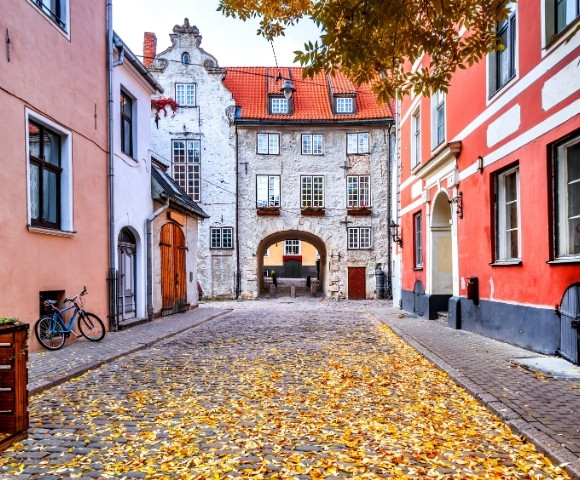A picturesque cobblestone street lined with colorful buildings in autumn. Leaves scatter the ground, and a bicycle rests on the left. A charming archway leads to more buildings beyond.