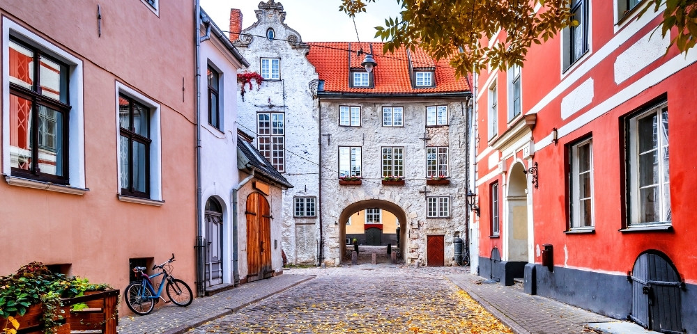 A picturesque cobblestone street lined with colorful buildings in autumn. Leaves scatter the ground, and a bicycle rests on the left. A charming archway leads to more buildings beyond.