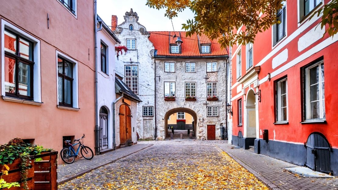 A picturesque cobblestone street lined with colorful buildings in autumn. Leaves scatter the ground, and a bicycle rests on the left. A charming archway leads to more buildings beyond.