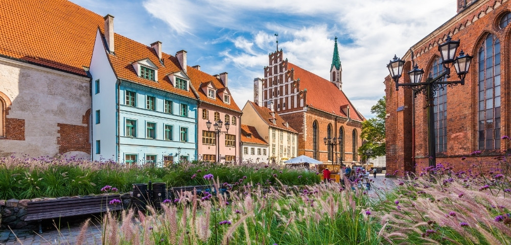 Historic European cityscape with colorful, gabled buildings and a brick church under a vivid blue sky. Ornamental grasses and benches in the foreground.