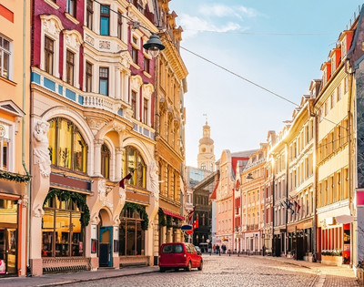 Charming European street with colorful historic buildings illuminated by warm sunlight. A red car and a few pedestrians on the cobblestone road convey a peaceful morning atmosphere.