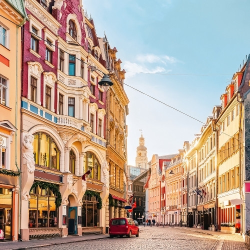 Charming European street with colorful historic buildings illuminated by warm sunlight. A red car and a few pedestrians on the cobblestone road convey a peaceful morning atmosphere.