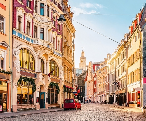Charming European street with colorful historic buildings illuminated by warm sunlight. A red car and a few pedestrians on the cobblestone road convey a peaceful morning atmosphere.